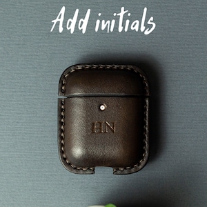 May include: A dark brown leather case for wireless earbuds featuring visible stitching along the edges and the embossed initials HN on the front. The text Add initials is printed in white above the case, which has a small circular cutout.