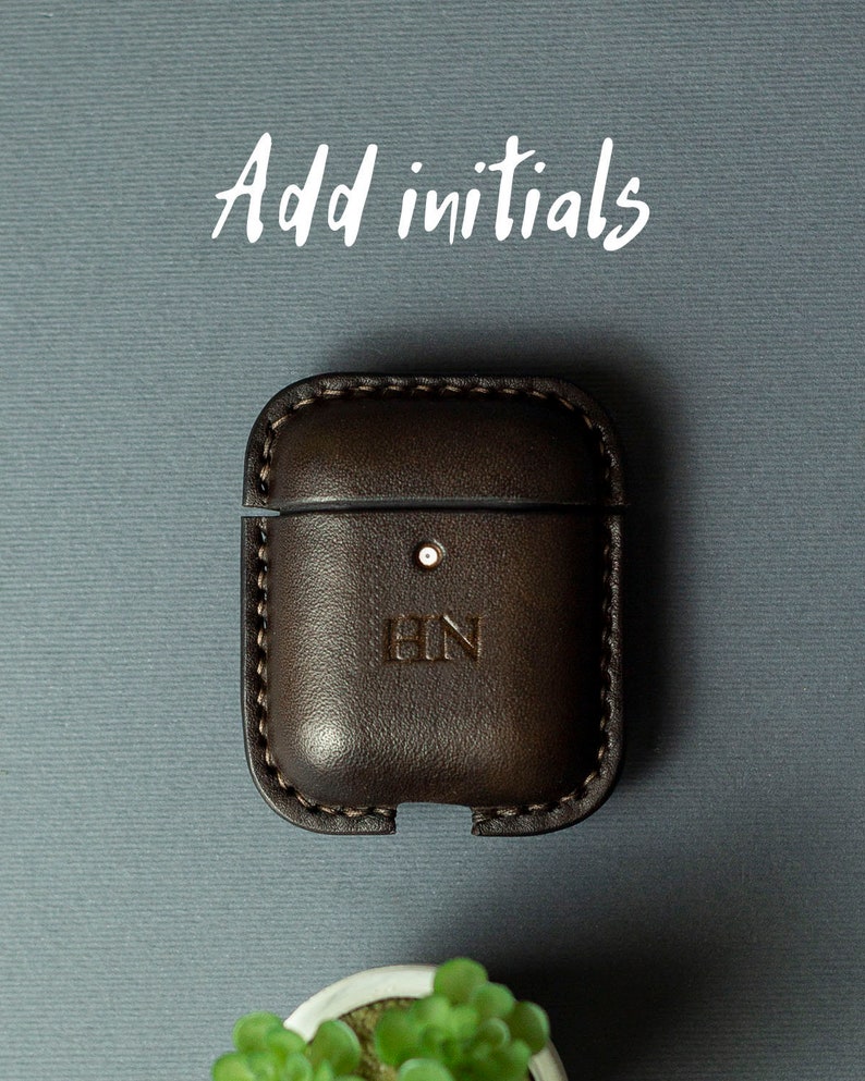 May include: A dark brown leather case for wireless earbuds featuring visible stitching along the edges and the embossed initials HN on the front. The text Add initials is printed in white above the case, which has a small circular cutout.