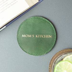 May include: A circular forest green leather coaster featuring visible perimeter stitching and the gold embossed text MOM'S KITCHEN centered on the surface. The coaster has a smooth matte finish and a consistent round shape with a clean edge design.
