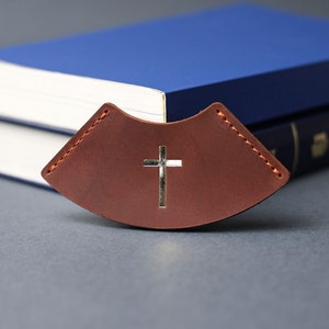 May include: A brown leather bookmark with a silver cross design. The bookmark is curved and has stitching around the edges.