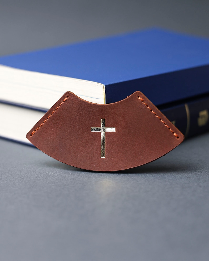 May include: A brown leather bookmark with a silver cross design. The bookmark is curved and has stitching around the edges.