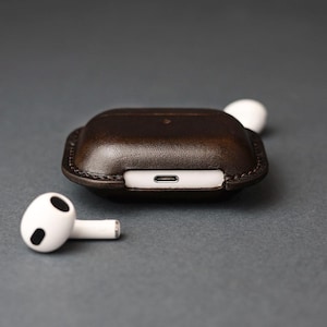 May include: A dark brown leather protective case for wireless earbuds, featuring visible stitching along the edges and a cutout for a charging port. Two white wireless earbuds are positioned near the case, one resting beside it and one partially visible.