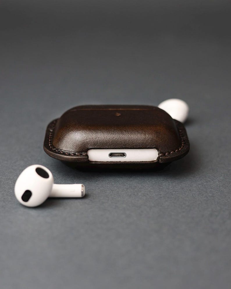 May include: A dark brown leather protective case for wireless earbuds, featuring visible stitching along the edges and a cutout for a charging port. Two white wireless earbuds are positioned near the case, one resting beside it and one partially visible.