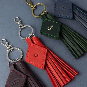 May include: A selection of leather keyrings with tassel detailing. Each keyring has a diamond-shaped tag with initials, in colours including red, green, blue, and burgundy. The keyrings have metal rings and clasps for attaching to keys or bags.