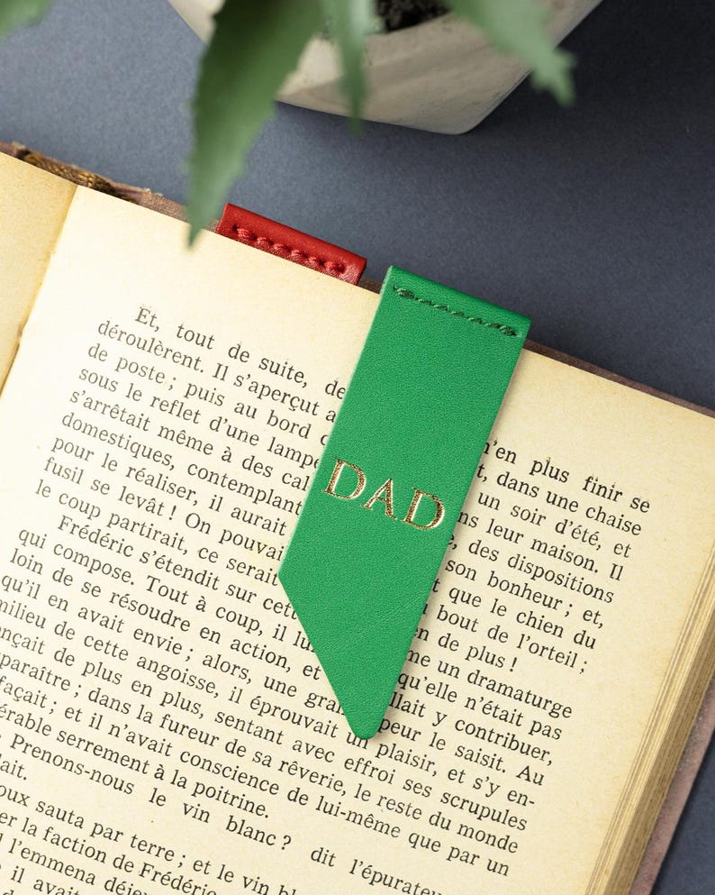 May include: Green leather bookmark with the gold embossed word "DAD". The bookmark is inserted into a book with printed text. A red leather bookmark is also visible.