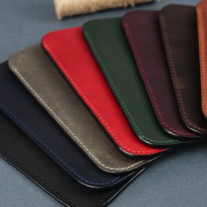 May include: A set of eight leather phone cases in various colors, including black, navy blue, gray, red, green, brown, burgundy, and orange. The cases are arranged in a fan shape, with the black case at the bottom and the orange case at the top. Each case has a stitched edge.