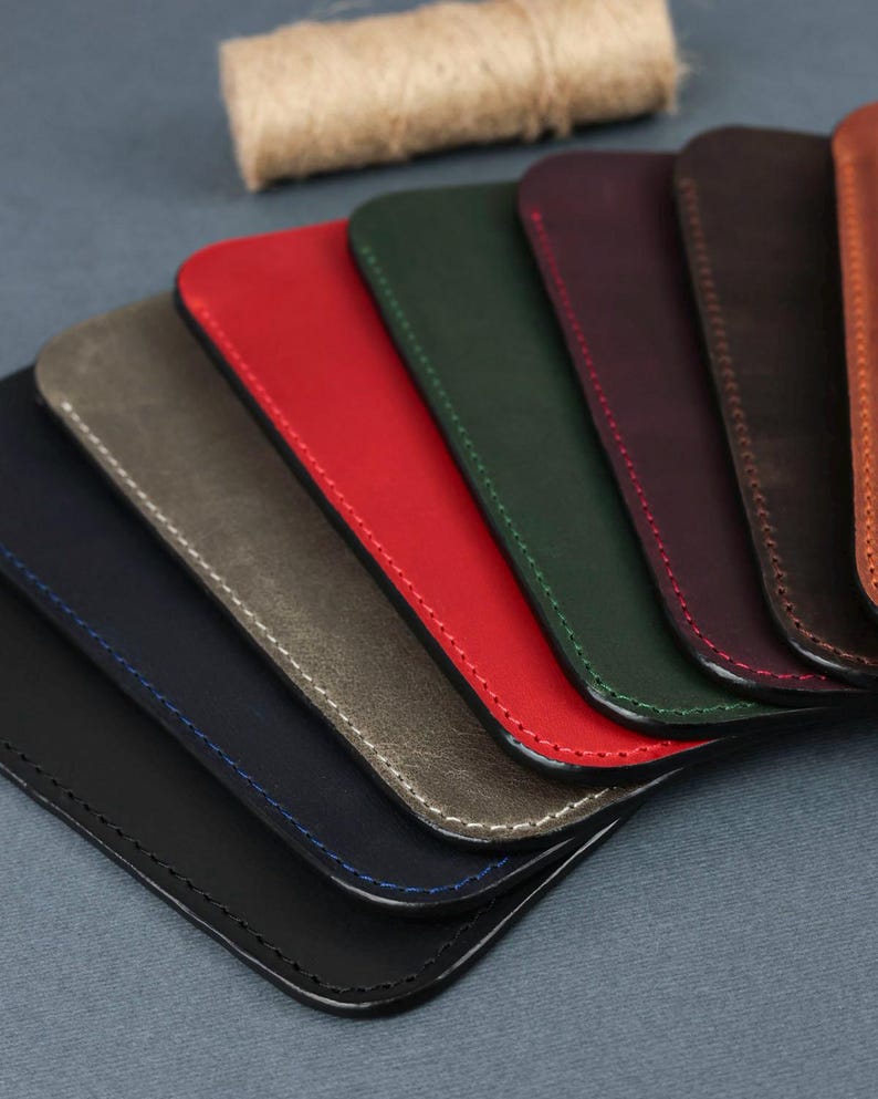 May include: A set of eight leather phone cases in various colors, including black, navy blue, gray, red, green, brown, burgundy, and orange. The cases are arranged in a fan shape, with the black case at the bottom and the orange case at the top. Each case has a stitched edge.