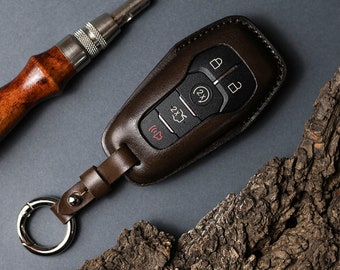 May include: A dark brown leather key fob cover encasing a black car key remote with five buttons, including lock, unlock, remote start, and panic icons. The cover features a matching leather strap attached to a silver metal circular keyring.