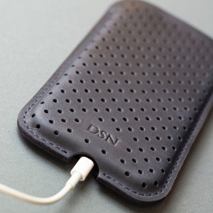 May include: Dark gray leather phone case with a perforated design and the embossed letters "DSN". A white charging cable is plugged into the bottom of the case. The case has blue stitching around the edges.