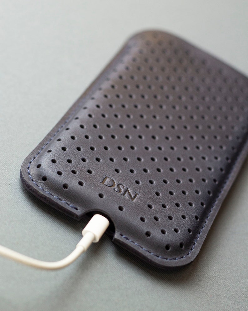 May include: Dark gray leather phone case with a perforated design and the embossed letters "DSN". A white charging cable is plugged into the bottom of the case. The case has blue stitching around the edges.