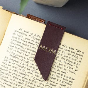 May include: A burgundy leather bookmark with the gold word "MOM" is in a book. The bookmark has a pointed end and is stitched with red thread. The book's pages are yellowed. The bookmark is a gift.