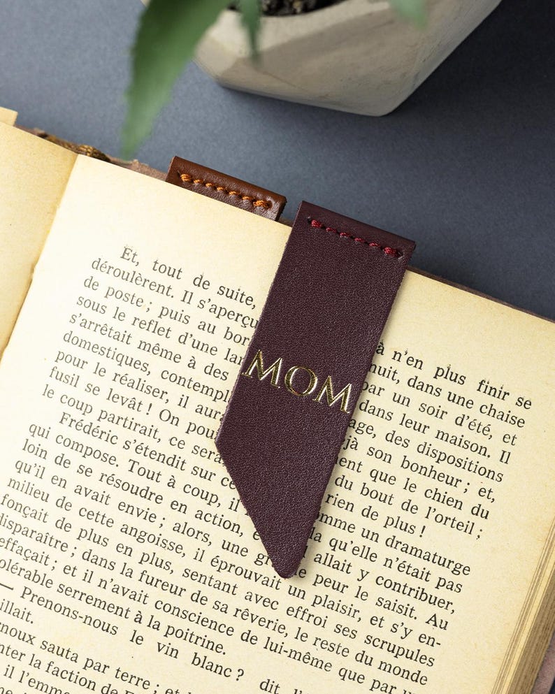 May include: A burgundy leather bookmark with the gold word "MOM" is in a book. The bookmark has a pointed end and is stitched with red thread. The book's pages are yellowed. The bookmark is a gift.