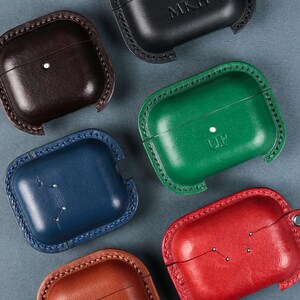 May include: Six leather cases for wireless earbuds in black, dark brown, green, blue, red, and tan. Each case features visible stitching along the edges, a small metal rivet, and some are embossed with initials like MKH, UP, or DM.