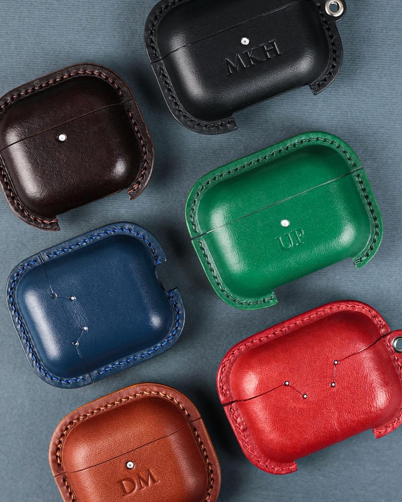 May include: Six leather cases for wireless earbuds in black, dark brown, green, blue, red, and tan. Each case features visible stitching along the edges, a small metal rivet, and some are embossed with initials like MKH, UP, or DM.