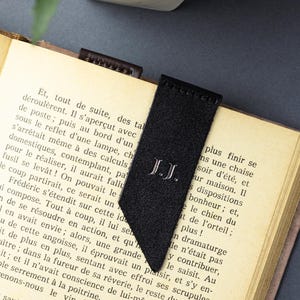 May include: A black suede bookmark with the initials "J.J." embroidered in white. The bookmark is inserted into a book with aged pages. The bookmark has a distinctive angled cut at the bottom.