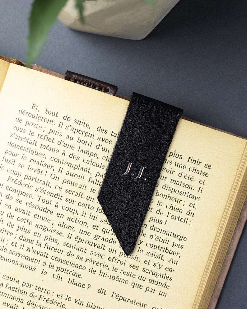 May include: A black suede bookmark with the initials "J.J." embroidered in white. The bookmark is inserted into a book with aged pages. The bookmark has a distinctive angled cut at the bottom.