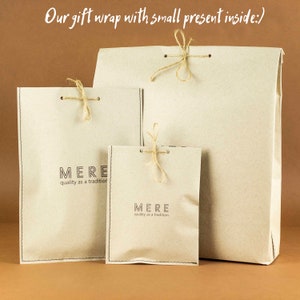May include: Three brown paper gift bags with twine ties. The bags are nested inside each other, with the largest bag on the bottom and the smallest bag on top. The bags have the text "MERE quality as a tradition" printed on them.