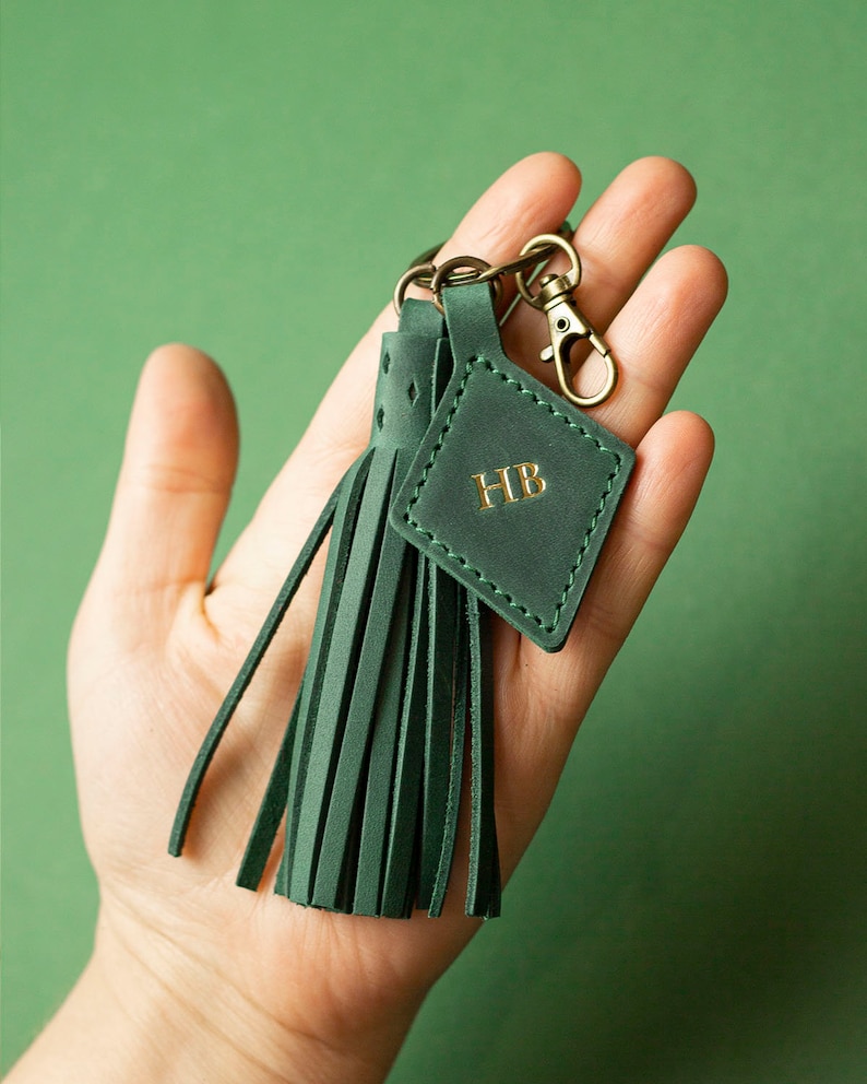 May include: A dark green leather keychain featuring a tassel and a square tag. The tag is embossed with the gold letters "HB" and is attached to a metal clasp. The keychain is held in a hand against a green backdrop.