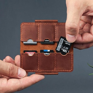 以下が含まれることがあります： 茶色の革製SDカードホルダー。複数のスロットがあり、開くと様々なSDカードが見え、黒いLexar Professional 128GBカードを挿入する手が見えます。このホルダーは、メモリーカードを保護し整理するように設計されています。