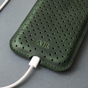 May include: A dark green leather phone case with a perforated design and the initials "AJR" embossed on the front. A white charging cable is plugged into the bottom of the case. The case is on a gray surface.