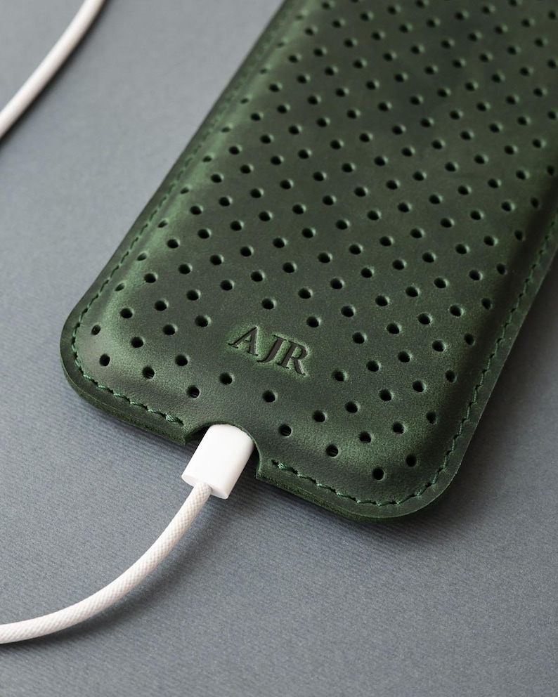 May include: A dark green leather phone case with a perforated design and the initials "AJR" embossed on the front. A white charging cable is plugged into the bottom of the case. The case is on a gray surface.