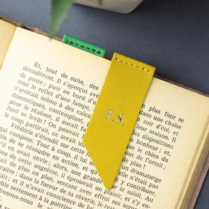 May include: A yellow leather bookmark with the initials "F.S." in silver lettering. The bookmark is inserted into a book with a green bookmark visible. The book is open to a page of text.