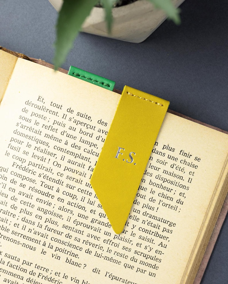 May include: A yellow leather bookmark with the initials "F.S." in silver lettering. The bookmark is inserted into a book with a green bookmark visible. The book is open to a page of text.