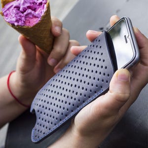 May include: A gray phone case with a perforated design and blue stitching holds a silver smartphone. A person holds the phone case in one hand and a purple ice cream cone in the other.