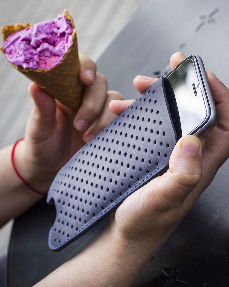 May include: A gray phone case with a perforated design and blue stitching holds a silver smartphone. A person holds the phone case in one hand and a purple ice cream cone in the other.