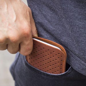 May include: A brown leather phone case with a perforated design is partially visible in a pocket. The case has a smooth, rounded edge and a series of small, evenly spaced holes. The case is being placed into a dark blue pocket.
