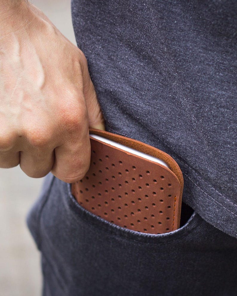 May include: A brown leather phone case with a perforated design is partially visible in a pocket. The case has a smooth, rounded edge and a series of small, evenly spaced holes. The case is being placed into a dark blue pocket.