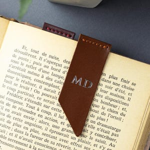 May include: A brown leather bookmark, embossed with the initials "MD" in silver, is placed in a book. A smaller, burgundy bookmark is also visible. The book has aged pages.