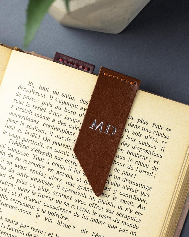 May include: A brown leather bookmark, embossed with the initials "MD" in silver, is placed in a book. A smaller, burgundy bookmark is also visible. The book has aged pages.
