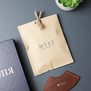 May include: A brown leather bookmark with the word "MERE" embossed in gold lettering. The bookmark is shaped like a crescent moon and has a stitched edge. The bookmark is packaged in a brown paper bag with the text "MERE FOUNDED IN UKRAINE IN 2012" printed on it.