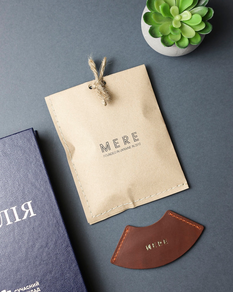 May include: A brown leather bookmark with the word "MERE" embossed in gold lettering. The bookmark is shaped like a crescent moon and has a stitched edge. The bookmark is packaged in a brown paper bag with the text "MERE FOUNDED IN UKRAINE IN 2012" printed on it.