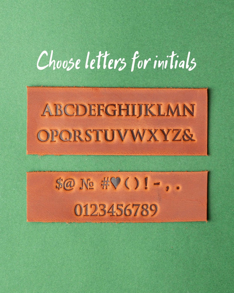 May include: Two rectangular brown leather stamps with embossed letters, numbers, and symbols. The top stamp displays the alphabet, and the bottom stamp shows numbers and symbols. The text "Choose letters for initials" is above the stamps on a green background.