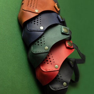 May include: Four leather face masks in different colors: brown, navy blue, green, and red. The masks have a perforated design and are stacked on top of each other.