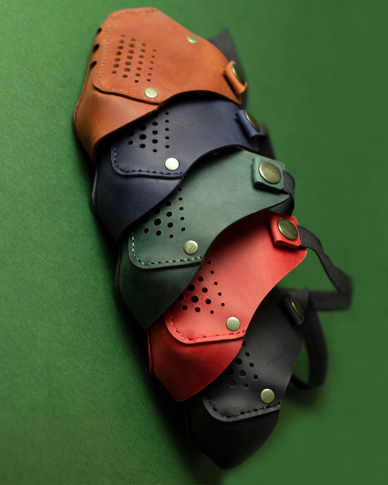 May include: Four leather face masks in different colors: brown, navy blue, green, and red. The masks have a perforated design and are stacked on top of each other.