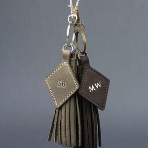 May include: Two leather keychains with diamond-shaped tags and fringe tassels. One is grey with "SB" initials, the other is brown with "MW" initials. Both have metal clasps and key rings, hanging against a grey background.