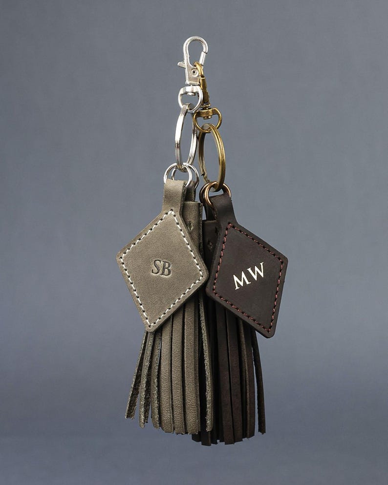 May include: Two leather keychains with diamond-shaped tags and fringe tassels. One is grey with "SB" initials, the other is brown with "MW" initials. Both have metal clasps and key rings, hanging against a grey background.