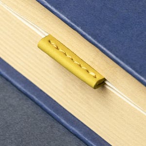 May include: A blue book with a yellow leather bookmark. The bookmark is stitched with yellow thread and is inserted between the pages. The pages are light beige.