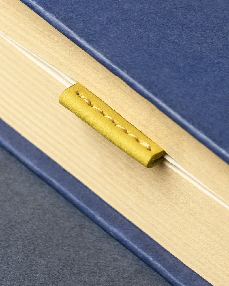May include: A blue book with a yellow leather bookmark. The bookmark is stitched with yellow thread and is inserted between the pages. The pages are light beige.