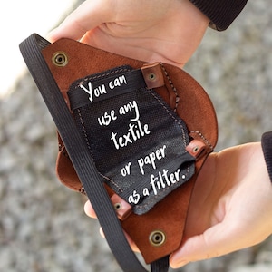 May include: A brown leather face mask with a black elastic strap. The mask has a pocket for a filter. The text "You can use any textile or paper as a filter." is written on the mask.
