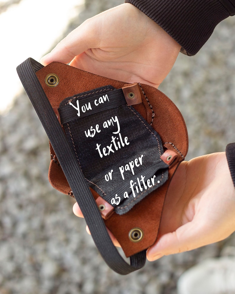 May include: A brown leather face mask with a black elastic strap. The mask has a pocket for a filter. The text "You can use any textile or paper as a filter." is written on the mask.