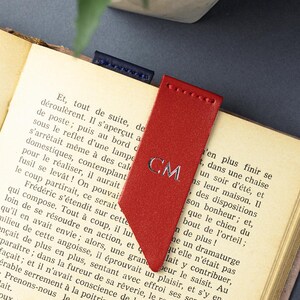 May include: A red leather bookmark with silver initials "CM". The bookmark is in a book with old pages. A small blue leather bookmark is also visible. The book is open.