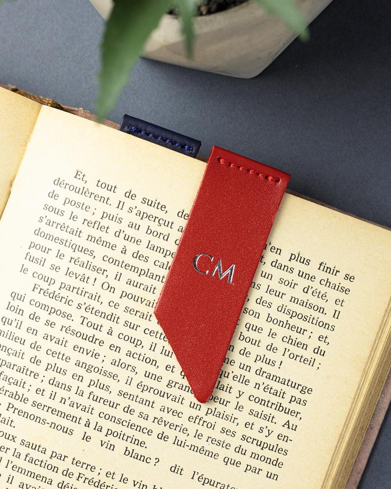 May include: A red leather bookmark with silver initials "CM". The bookmark is in a book with old pages. A small blue leather bookmark is also visible. The book is open.