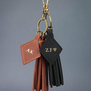 May include: Three personalised leather keyrings. One is brown with "AK" initials, another is black with "Z.F &hearts;", and the third is brown. Each keyring has a tassel and is attached to a gold-coloured clasp and ring.
