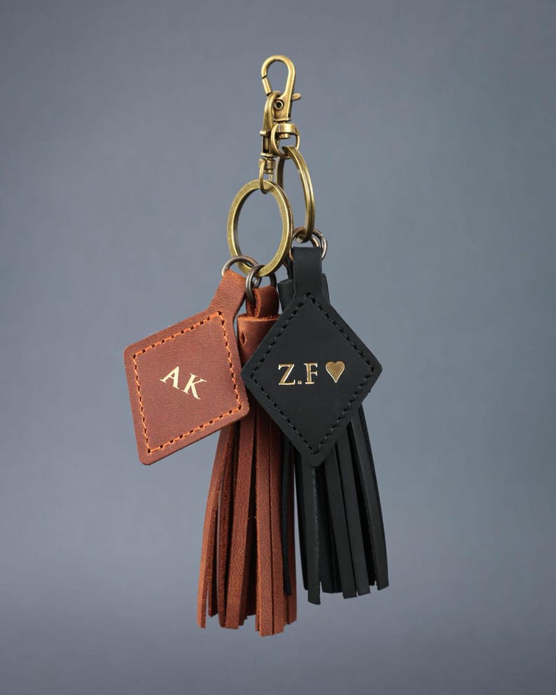 May include: Three personalised leather keyrings. One is brown with "AK" initials, another is black with "Z.F &hearts;", and the third is brown. Each keyring has a tassel and is attached to a gold-coloured clasp and ring.