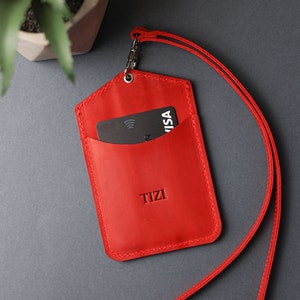 May include: Red leather ID badge holder with a black card inside and a red lanyard. The holder has the word "TIZI" embossed on the front.