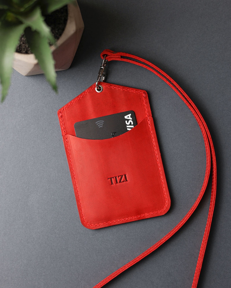 May include: Red leather ID badge holder with a black card inside and a red lanyard. The holder has the word "TIZI" embossed on the front.
